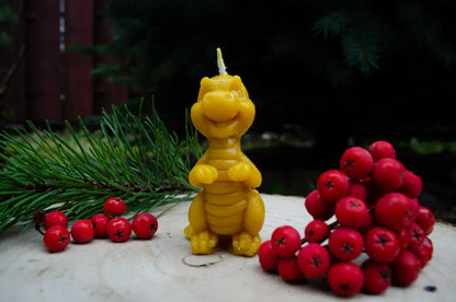 Natūralaus bičių vaško žvakė drakoniuko formos pastatyta ant kelmo šalia pušies ir eglių.