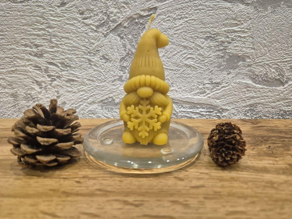 Kalėdinė bičių vaško žvakė elfas su snaige ant stiklinio padėkliuko šalia kankorėžių.