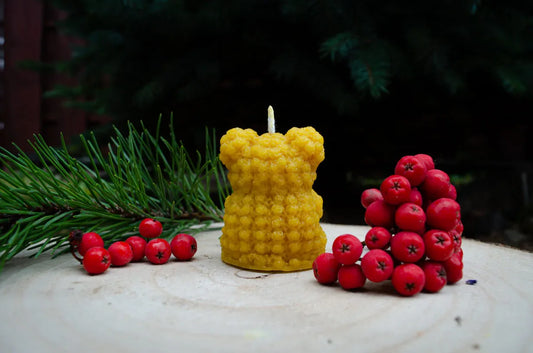 Rankų darbo bičių vaško žvakė – meškiukas su širdele, natūralaus geltono vaško figūrėlė ant medinio kelmo atsukta nugara į žiūrova.